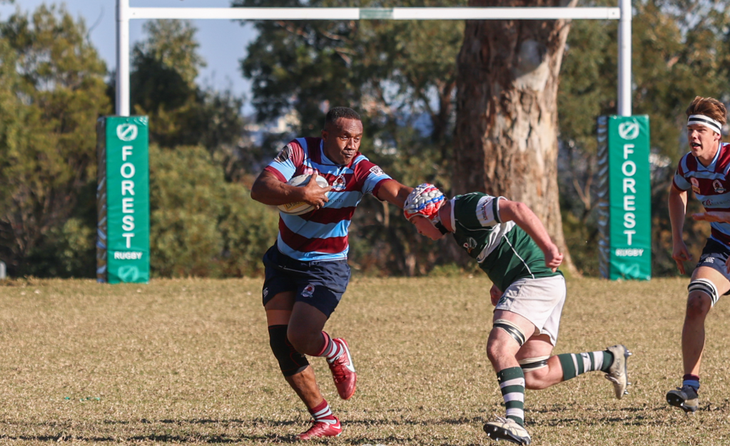A rugby player in a blue and maroon striped jersey runs with the ball, evading a tackle from an opponent in a green and white striped jersey. In the background, rugby goalposts and sidelines are visible.