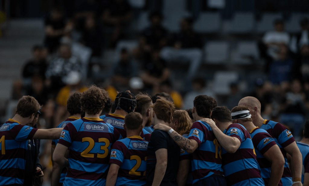 A rugby team huddled together in a circle, wearing striped jerseys in blue and maroon, preparing for an upcoming match.