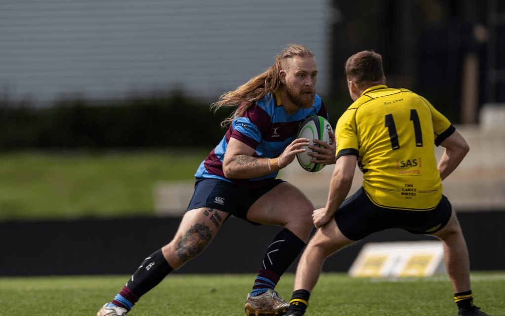 A rugby player with long hair and tattoos is holding a rugby ball, facing an opponent in a yellow jersey with the number 11. The scene takes place on a grass field.