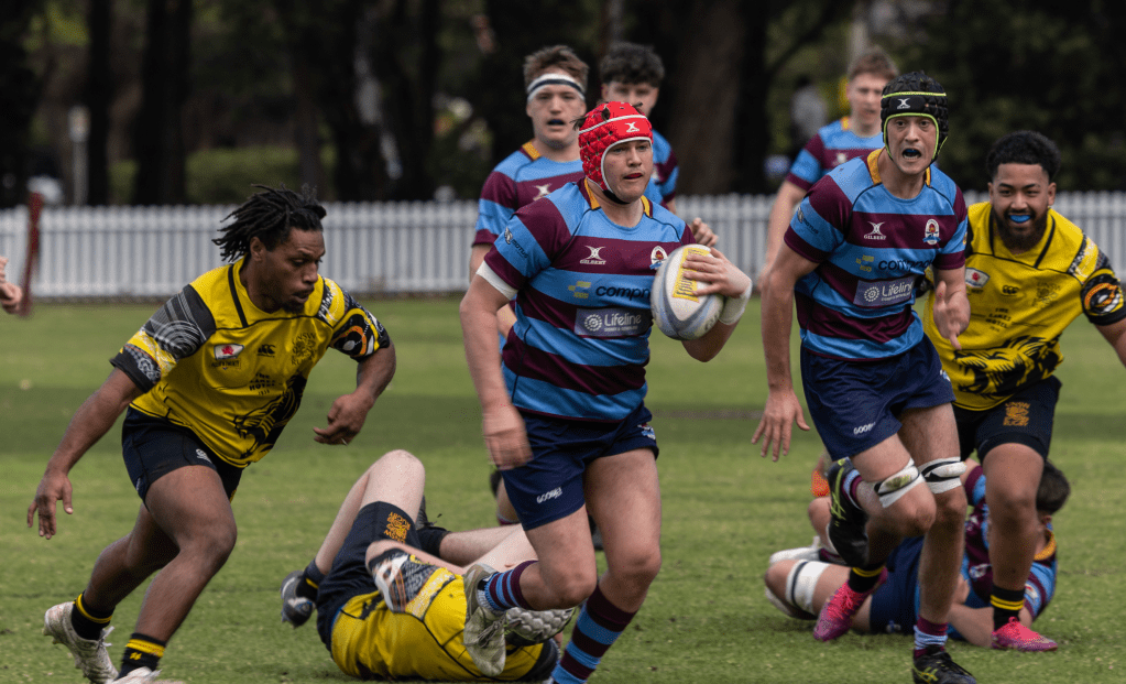 A rugby match in progress with players in action. A player in maroon and blue stripes runs with the ball while others are in pursuit. Various players in yellow are also engaged in the play, with one player on the ground.